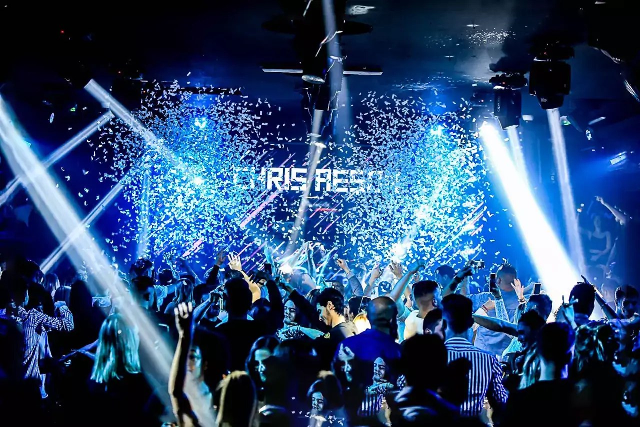 Foule en discothèque les bras levés sous confettis et faisceaux lumineux bleus